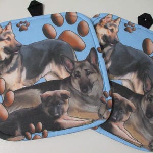 May include: Two blue pot holders with a paw print design. Each pot holder features a German Shepherd dog and a puppy.