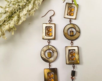 May include: A pair of long, dangling earrings with a geometric design. The earrings are made of brown and white shell-like material with amber-colored beads. The earrings have a square, circle, and another square shape.