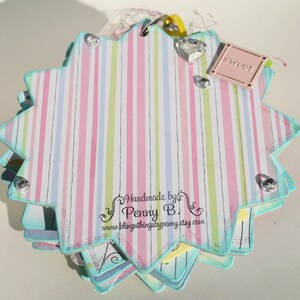 May include: A multi-layered, star-shaped scrapbook with a pink, green, and blue striped cover. The cover has a small pink tag with the word "sweet" on it. The scrapbook is decorated with rhinestones and has a handwritten label that reads "Handmade by Penny B. www.blingthingsbypenny.etsy.com".