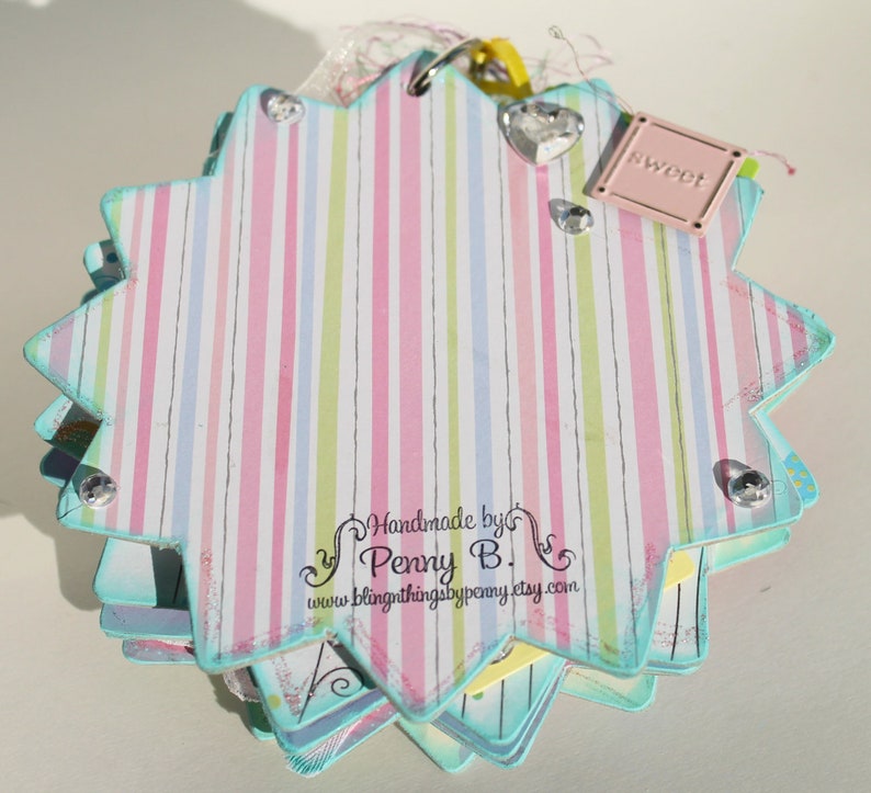May include: A multi-layered, star-shaped scrapbook with a pink, green, and blue striped cover. The cover has a small pink tag with the word "sweet" on it. The scrapbook is decorated with rhinestones and has a handwritten label that reads "Handmade by Penny B. www.blingthingsbypenny.etsy.com".