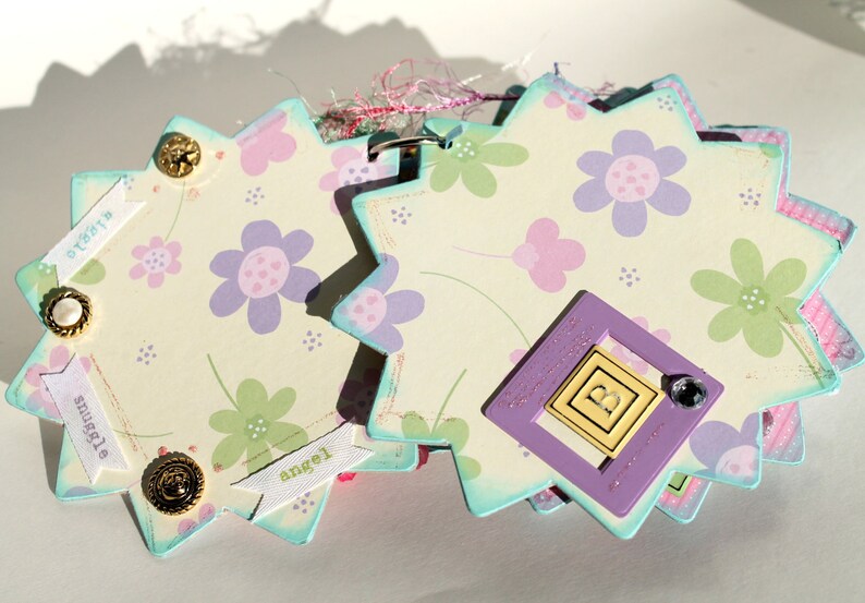 May include: A multi-layered scrapbook page with a light blue, white, and pink floral design. The page has a purple frame with the letter "B" inside. The page also has several white banners with the words "sweetest", "snuggle", and "angel" written on them.