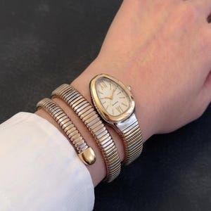 May include: A gold and silver-toned quartz watch with a white face and gold hands is worn on a wrist, accompanied by a matching gold and silver bracelet. The watch has an oval face and a flexible band. The bracelet wraps around the wrist.