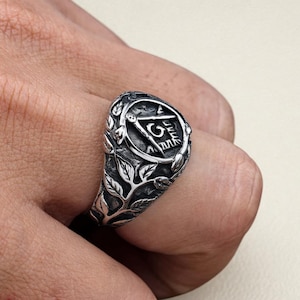 May include: Silver-toned ring featuring a Masonic symbol within an oval frame, surrounded by detailed leaf and vine designs. The ring's intricate details and polished finish create a classic, elegant look.