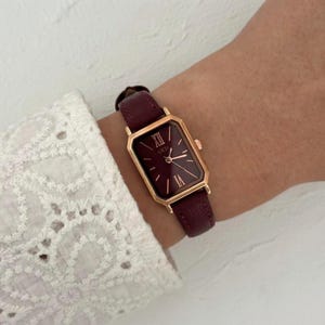 May include: A burgundy rectangular wristwatch with a gold-colored frame and a matching burgundy leather band. The watch face displays Roman numerals and the brand name "CEDI". The watch is worn on a wrist.