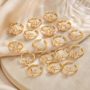 Op de afbeelding: Een collectie verstelbare goudkleurige ringen, elk voorzien van een letter van het alfabet versierd met kleine witte kunstparels. De ringen hebben een open ontwerp voor maatverstelling en zijn gerangschikt op een glad oppervlak.