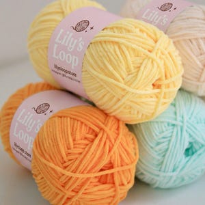 May include: A collection of colorful yarn balls in shades of yellow, orange, and light blue. Each ball is wrapped with a pink label that reads "Lily's Loop" and includes the Instagram handle "lilysloop.store". The yarn is ideal for knitting and crochet projects.