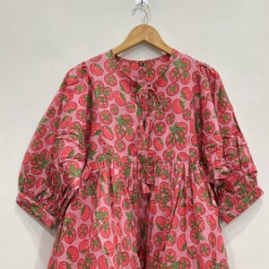 May include: A pink dress with a repeating pattern of red tomatoes and green leaves. The dress has a round neckline with a tie, short sleeves, and a gathered waist. The dress is hanging on a wooden hanger.