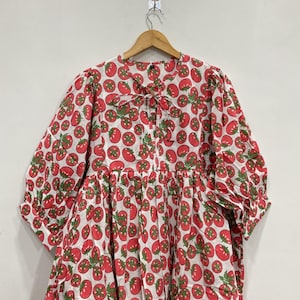 May include: A white dress with a red tomato pattern and green leaves. The dress has a round neckline, long sleeves, and a gathered skirt. The dress is hanging on a wooden hanger.