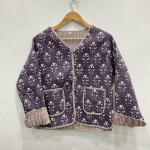 May include: A quilted jacket in a deep purple hue featuring a white floral block print pattern. The garment has a V-neckline, long sleeves with striped cuffs, and two front patch pockets. The interior and edges are lined with a brown and white striped fabric.