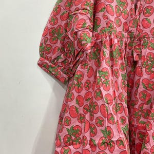 May include: A pink dress with a repeating pattern of red tomatoes and green leaves. The dress has short sleeves and a gathered waist. The fabric appears to be cotton or a similar lightweight material.