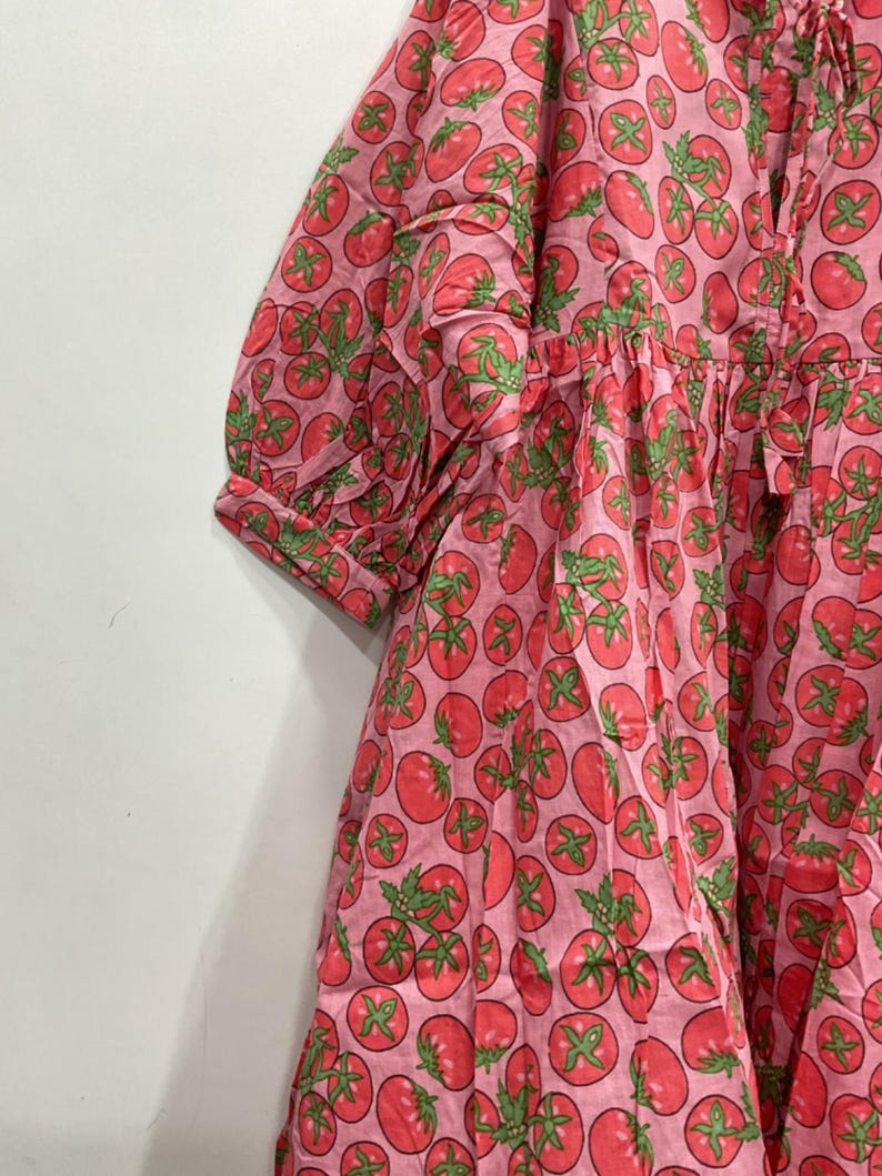 May include: A pink dress with a repeating pattern of red tomatoes and green leaves. The dress has short sleeves and a gathered waist. The fabric appears to be cotton or a similar lightweight material.