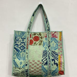 May include: A patchwork tote bag in various patterns and colours, including blue, red, and green. The bag has a light green background and green striped trim and handles. The bag is hanging on a white wall.