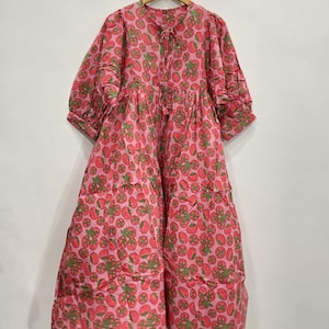 May include: A pink dress with a repeating pattern of red tomatoes and green leaves. The dress has a round neckline, short sleeves, and a tiered skirt. It is hanging on a wooden hanger.
