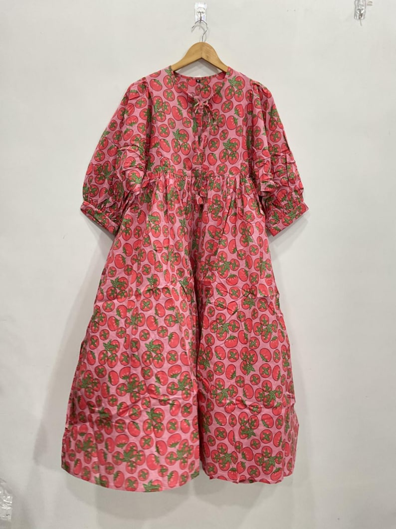 May include: A pink dress with a repeating pattern of red tomatoes and green leaves. The dress has a round neckline, short sleeves, and a tiered skirt. It is hanging on a wooden hanger.
