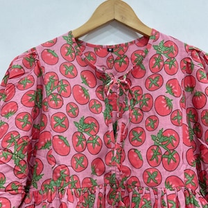 May include: A pink dress with a repeating pattern of red tomatoes and green leaves. The dress has a round neckline with a tie closure, short puffed sleeves, and a gathered skirt. The fabric appears to be cotton.