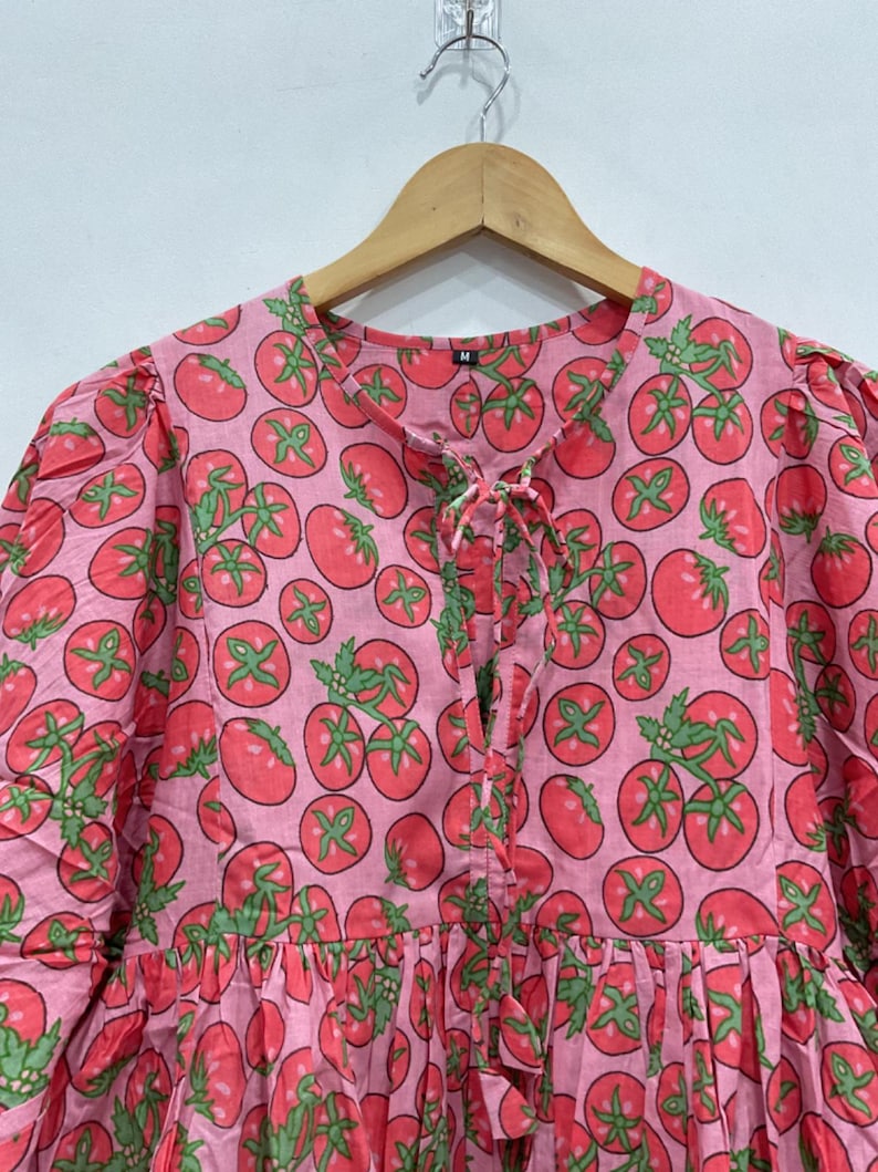 May include: A pink dress with a repeating pattern of red tomatoes and green leaves. The dress has a round neckline with a tie closure, short puffed sleeves, and a gathered skirt. The fabric appears to be cotton.