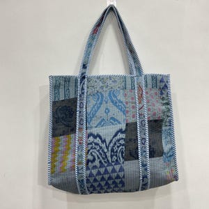 Puede incluir: Un bolso tote patchwork con una variedad de telas estampadas azules y grises. El bolso tiene forma rectangular con un borde a rayas y asas a juego. Cuelga contra una pared blanca.