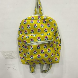 Puede incluir: Una mochila acolchada de color amarillo con un patrón repetitivo de pequeñas flores moradas con hojas verdes. La bolsa incluye un bolsillo frontal con cremallera, un asa superior y correas ajustables para los hombros, todo con un borde a rayas blancas y verdes.