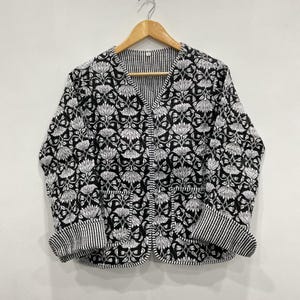 Könnte beinhalten: Schwarz-weiß bedruckte Jacke mit Blumenmuster, V-Ausschnitt und zwei Taschen. Die Jacke hat ein schwarz-weiß gestreiftes Futter und Manschetten. Die Jacke hängt an einem Holzbügel.