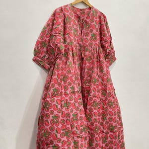 May include: A pink dress with a repeating pattern of red tomatoes and green leaves. The dress has a round neckline, long sleeves, and a tiered skirt. It is hanging on a wooden hanger against a white wall.