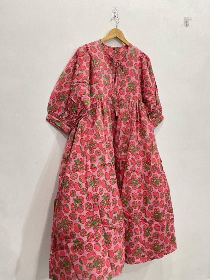 May include: A pink dress with a repeating pattern of red tomatoes and green leaves. The dress has a round neckline, long sleeves, and a tiered skirt. It is hanging on a wooden hanger against a white wall.