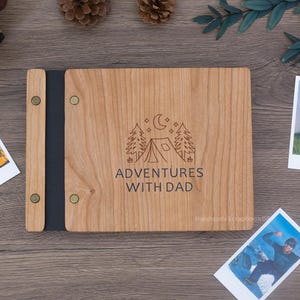 May include: A rectangular wooden photo album with a light brown grain finish. The cover features an engraved graphic of a tent, pine trees, and a crescent moon above the text ADVENTURES WITH DAD. It has a black spine secured by four brass rivets.