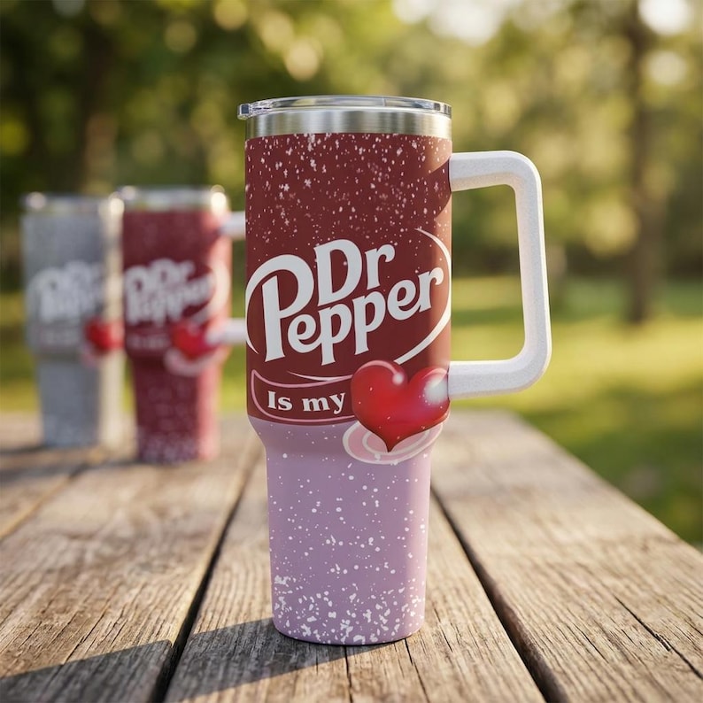 Puede incluir: Un vaso de acero inoxidable con asa y tapa blancas. El vaso presenta el logotipo de Dr Pepper con un coraz&oacute;n y la frase "Is my". El vaso tiene una parte superior burdeos y una inferior lavanda con motas blancas.