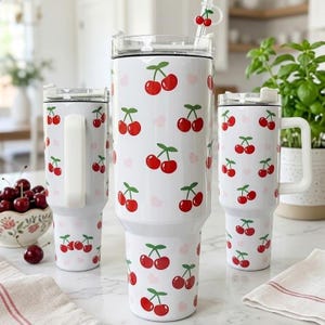 May include: White insulated tumblers with a red cherry pattern and pink hearts. The tumblers have clear lids and a white handle. One tumbler has a straw with a cherry charm. A bowl of cherries is in the background.