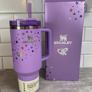 K&ouml;nnte beinhalten: Ein lavendelfarbener Stanley Quencher H2.0 Flowstate-Becher mit Griff und Strohhalm. Der Becher ist mit bunten Sternen und dem Stanley-Logo verziert. Die daneben stehende Box hat das gleiche Logo und Sterndesign.