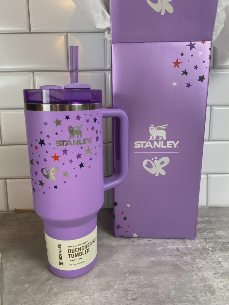K&ouml;nnte beinhalten: Ein lavendelfarbener Stanley Quencher H2.0 Flowstate-Becher mit Griff und Strohhalm. Der Becher ist mit bunten Sternen und dem Stanley-Logo verziert. Die daneben stehende Box hat das gleiche Logo und Sterndesign.