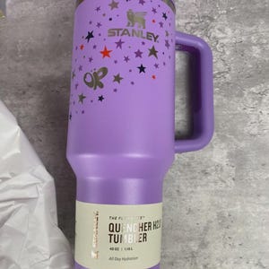 K&ouml;nnte beinhalten: Ein lila Stanley Quencher H2.0 Becher mit Henkel, verziert mit Sternen und Schmetterlingen. Der Becher hat einen Strohhalm und ein Etikett mit der Aufschrift "QUENCHER H2O TUMBLER" und fasst 1,18 L.