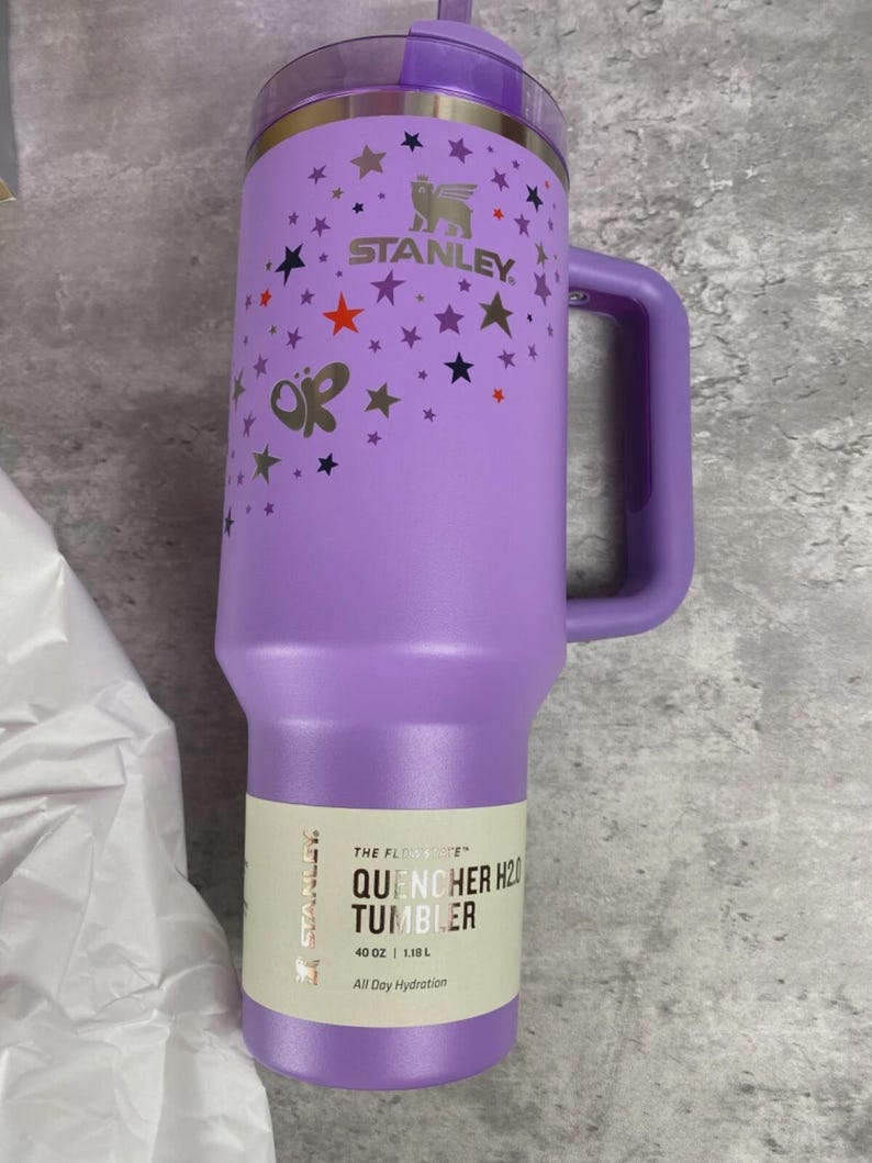 K&ouml;nnte beinhalten: Ein lila Stanley Quencher H2.0 Becher mit Henkel, verziert mit Sternen und Schmetterlingen. Der Becher hat einen Strohhalm und ein Etikett mit der Aufschrift "QUENCHER H2O TUMBLER" und fasst 1,18 L.