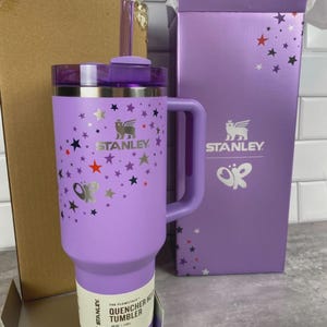 K&ouml;nnte beinhalten: Ein lila Stanley Quencher Hot Tumbler mit Griff, verziert mit Sternen. Der Becher hat ein Fassungsverm&ouml;gen von 1,18L. Die Box ist lila mit dem Stanley-Logo und Sterndesigns. Der Becher hat einen Strohhalm und einen Deckel.