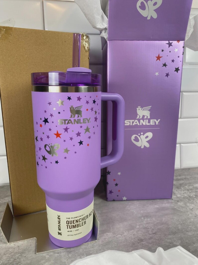 K&ouml;nnte beinhalten: Ein lila Stanley Quencher Hot Tumbler mit Griff, verziert mit Sternen. Der Becher hat ein Fassungsverm&ouml;gen von 1,18L. Die Box ist lila mit dem Stanley-Logo und Sterndesigns. Der Becher hat einen Strohhalm und einen Deckel.