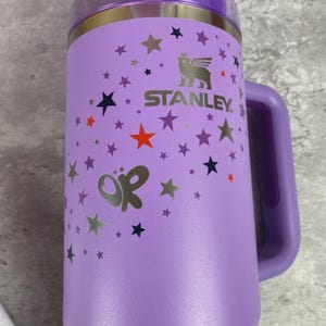 K&ouml;nnte beinhalten: Ein lila Stanley-Becher mit Griff, Deckel und Strohhalm. Der Becher ist mit silbernen Sternen und einem Schmetterlingsdesign verziert. Das Stanley-Logo ist in Silber sichtbar. F&uuml;r kalte Getr&auml;nke konzipiert.