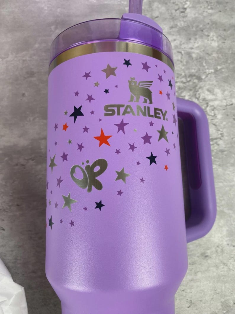 K&ouml;nnte beinhalten: Ein lila Stanley-Becher mit Griff, Deckel und Strohhalm. Der Becher ist mit silbernen Sternen und einem Schmetterlingsdesign verziert. Das Stanley-Logo ist in Silber sichtbar. F&uuml;r kalte Getr&auml;nke konzipiert.
