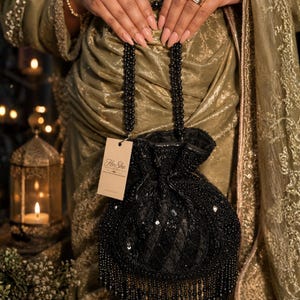 Könnte beinhalten: Eine schwarze Beuteltasche mit Kordelzug, verziert mit aufwendigen Perlenarbeiten, Pailletten und einem Rautenmuster. Die Tasche hat zwei Perlenhenkel und einen Saum aus hängenden schwarzen Perlen. Ein beiges Etikett mit dem Text HoShe Luxury ist befestigt.