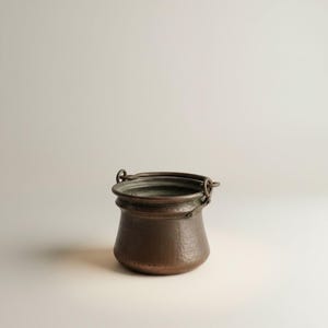 May include: A small, antique copper pot with a hammered texture. The pot has a rounded body and a flared rim, with a handle on each side. The pot is a dark, aged copper color.