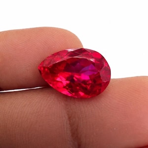 Könnte beinhalten: Ein leuchtend tiefroter Edelstein im Tropfenschliff mit zahlreichen Facetten, die das Licht reflektieren. Der Stein zeigt eine brillante, gesättigte karmesinrote Farbe auf seiner gesamten Oberfläche und weist eine klare, transparente Struktur auf.