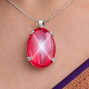 May include: A pendant necklace featuring a large, oval-shaped, vibrant red gemstone with a distinct six-rayed star pattern visible on its surface. The gemstone is held in a silver-toned metal setting with four prongs and hangs from a delicate chain.