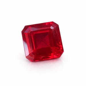 May include: A faceted, square-cut ruby gemstone. The gem is a deep, vibrant red color with multiple facets that reflect light. The gemstone is set against a white background.