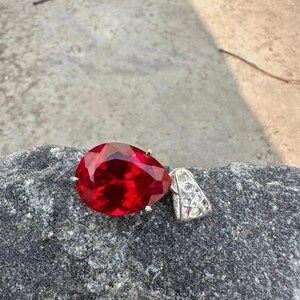 May include: A large, teardrop-shaped red gemstone pendant with a silver bail. The gemstone is faceted, catching the light and displaying a deep, rich color. The pendant is set against a gray, textured stone surface.