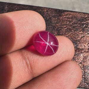 May include: A polished, oval-shaped, star ruby gemstone. The gem is a deep, rich red color with a six-rayed star pattern across its surface. The star effect is created by white, intersecting lines. The gemstone is held between fingers.