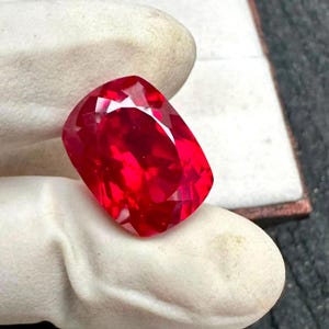 Pode incluir: Uma gema vermelha vibrante, lapidada em formato de almofada, com uma superfície altamente facetada que reflete a luz. A pedra exibe um tom carmesim profundo e saturado, mantida entre dois dedos cobertos por tecido branco.