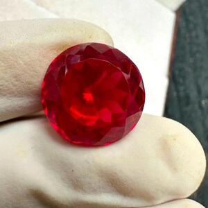 Pode incluir: Uma pedra preciosa redonda e facetada com uma cor vermelha vibrante e profunda. A pedra apresenta um corte brilhante com numerosas facetas triangulares e em forma de pipa que refletem a luz, criando um aspeto cintilante na superfície.