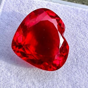 Può includere: Una pietra preziosa a forma di cuore di un colore rosso vivido e profondo con una superficie sfaccettata che riflette la luce. La pietra presenta un design simmetrico con una base appuntita e una parte superiore arrotondata e trasparente.