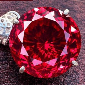 Pode incluir: Um pingente de pedra preciosa vermelho vibrante, redondo e facetado, montado em uma estrutura de metal prateado com garras. A pedra apresenta um corte brilhante com inúmeras facetas triangulares e a argola tem um padrão decorativo vazado.