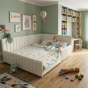 May include: A cream-colored upholstered corner daybed with vertical channel tufting, featuring a white duvet cover with a colorful space-themed pattern, positioned in a room with light green walls and light wood flooring.