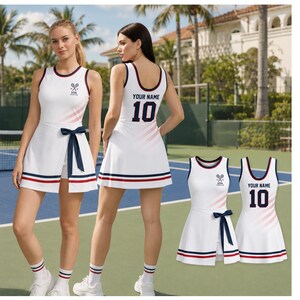 Może przedstawiać: Biała sukienka tenisowa z granatowo-czerwonym paskiem u dołu, granatowym wiązaniem w talii i małym logo USA Tennis Club na piersi. Z tyłu znajduje się napis YOUR NAME oraz numer 10 w kolorze granatowym, z subtelnym różowym gradientem.