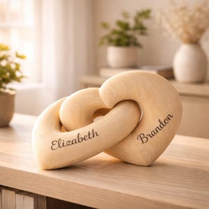 May include: Two interlocking wooden hearts, engraved with the names "Elizabeth" and "Brandon." The hearts are light-colored, possibly maple or birch, and sit on a wooden surface. The background includes plants and a vase.